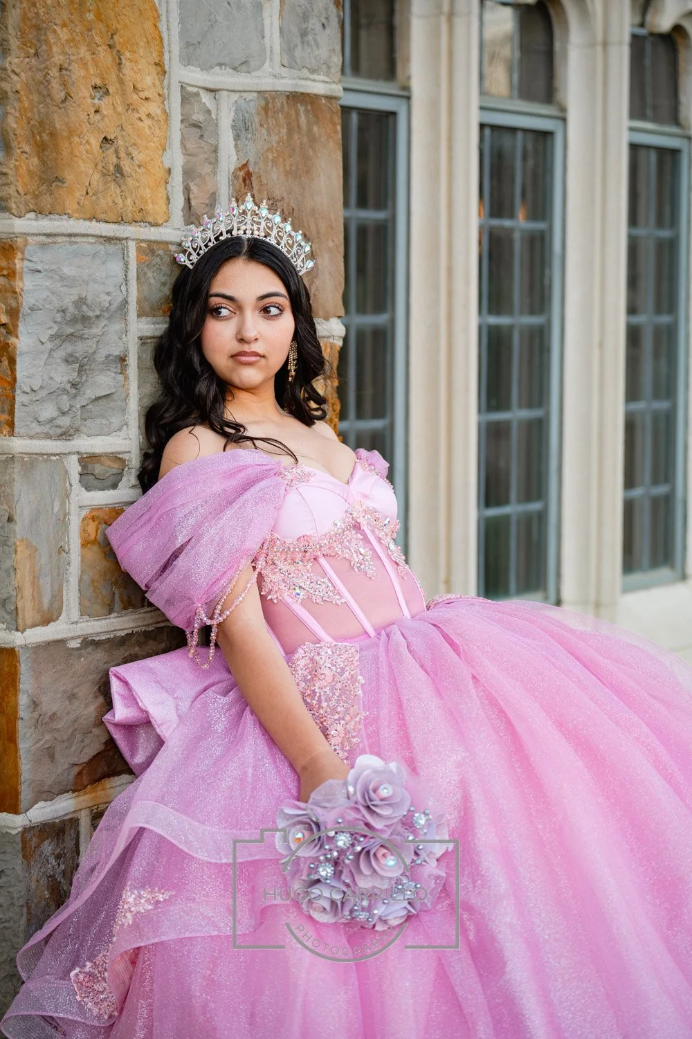 A quinceañera wearing a pink princess dress with off-shoulder puffed sleeves and a tiara, holding a bouquet of pink and purple flowers, sitting outdoors near a stone wall and large windows.