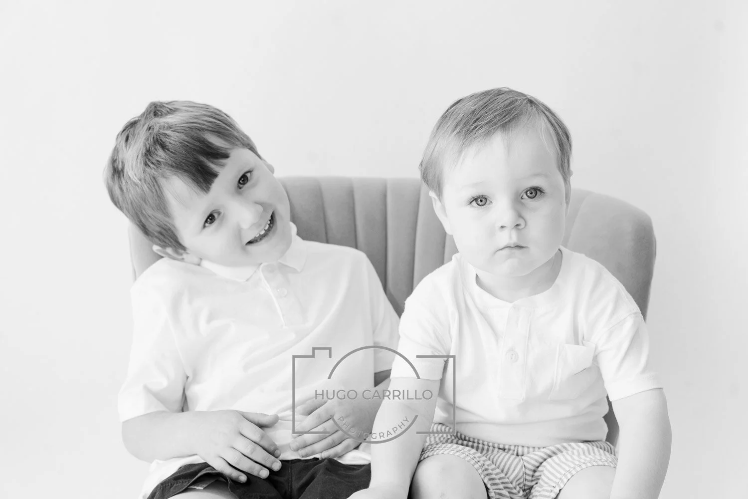 Two young boys sitting on a padded chair, one smiling with his head tilted, the other with a neutral expression, both wearing light-colored shirts.