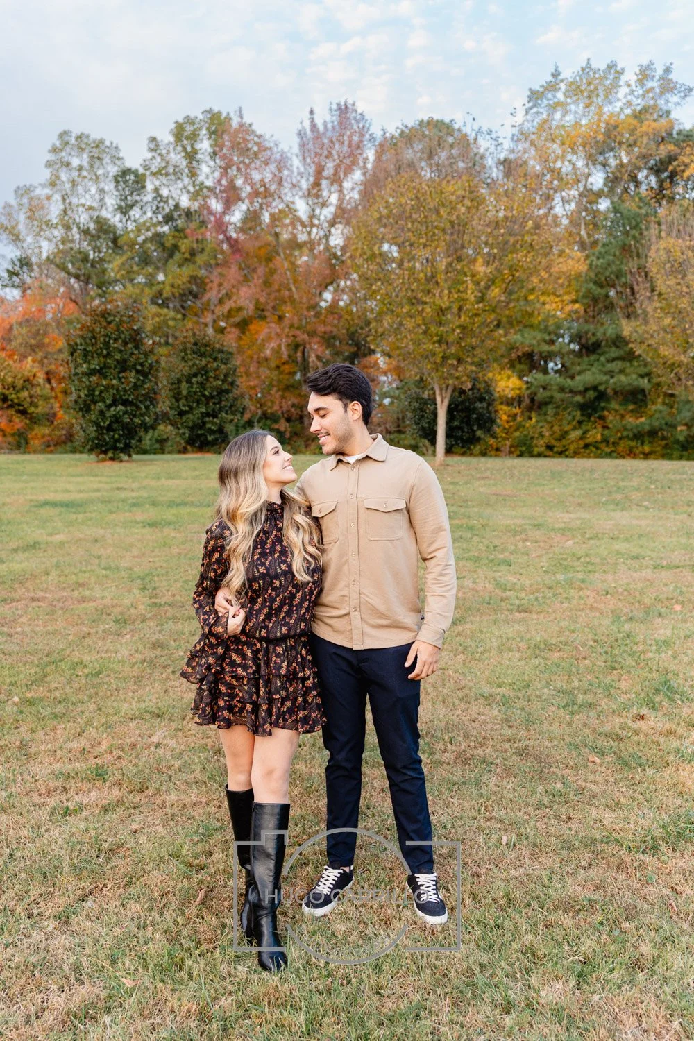 A young couple stands close together on a grassy field with autumn trees in the background. The woman wears a black floral dress and tall black boots, and the man wears a tan button-up shirt and dark pants. They smile at each other, sharing an intima