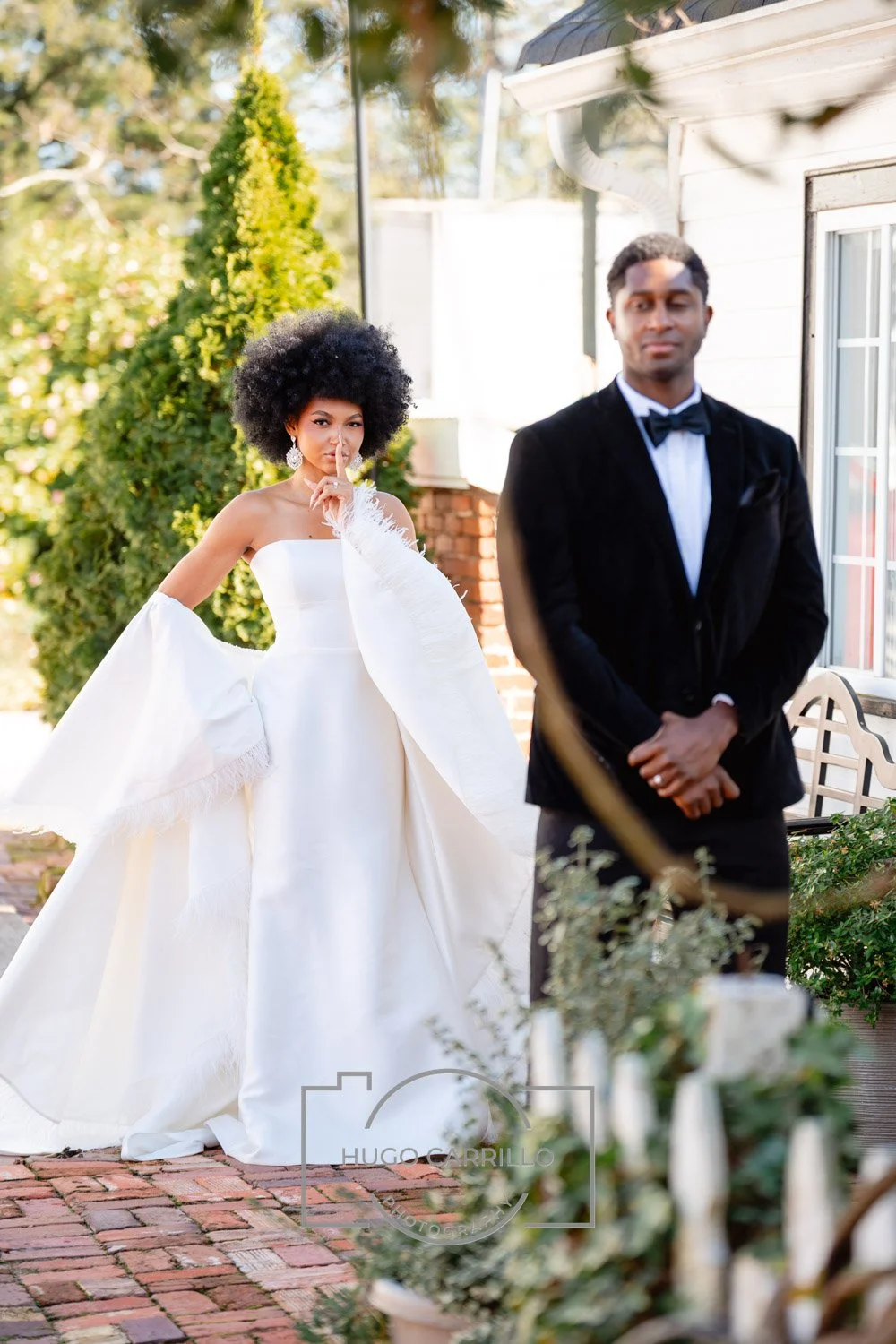 Bride with a big afro posing with her finger on her lips and wearing a white gown with dramatic sleeves, and groom in a black tuxedo with a bow tie standing with his hands clasped in front, outdoors near a white house with greenery.