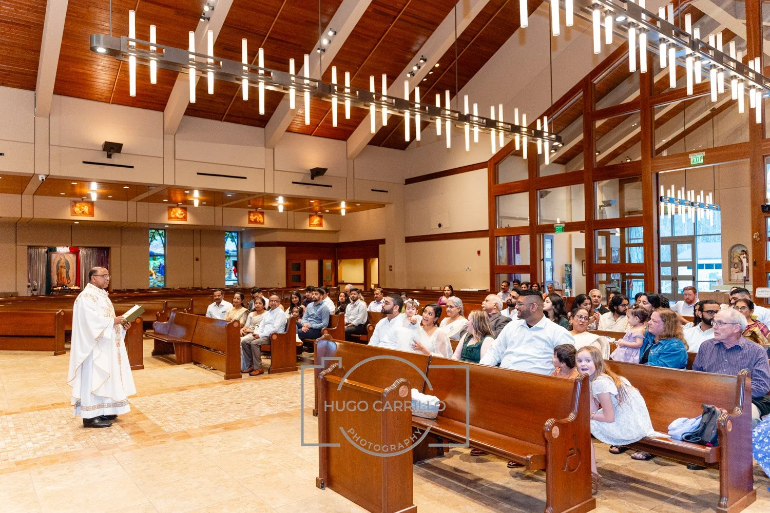 Catholic priest in white robes leading a service in a church with a congregation, wooden pews, and stained glass windows.