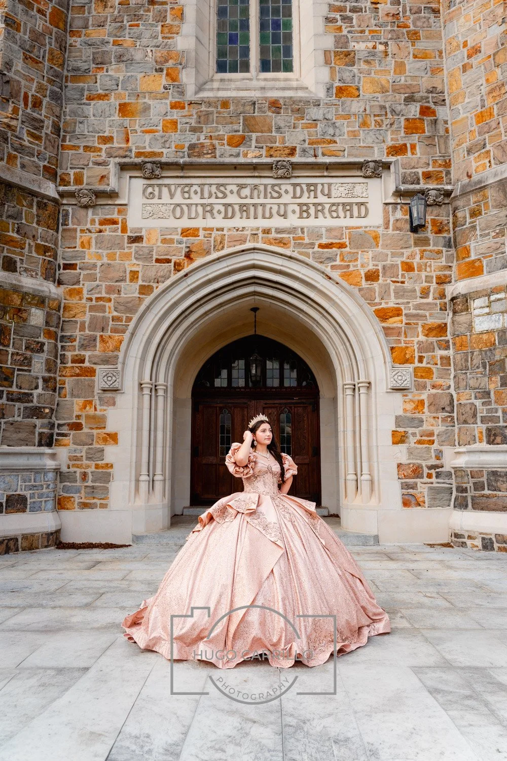 A quinceañera in a pink princess gown and crown standing in front of a stone church entrance with an inscription reading 'Give us this day our daily bread.'