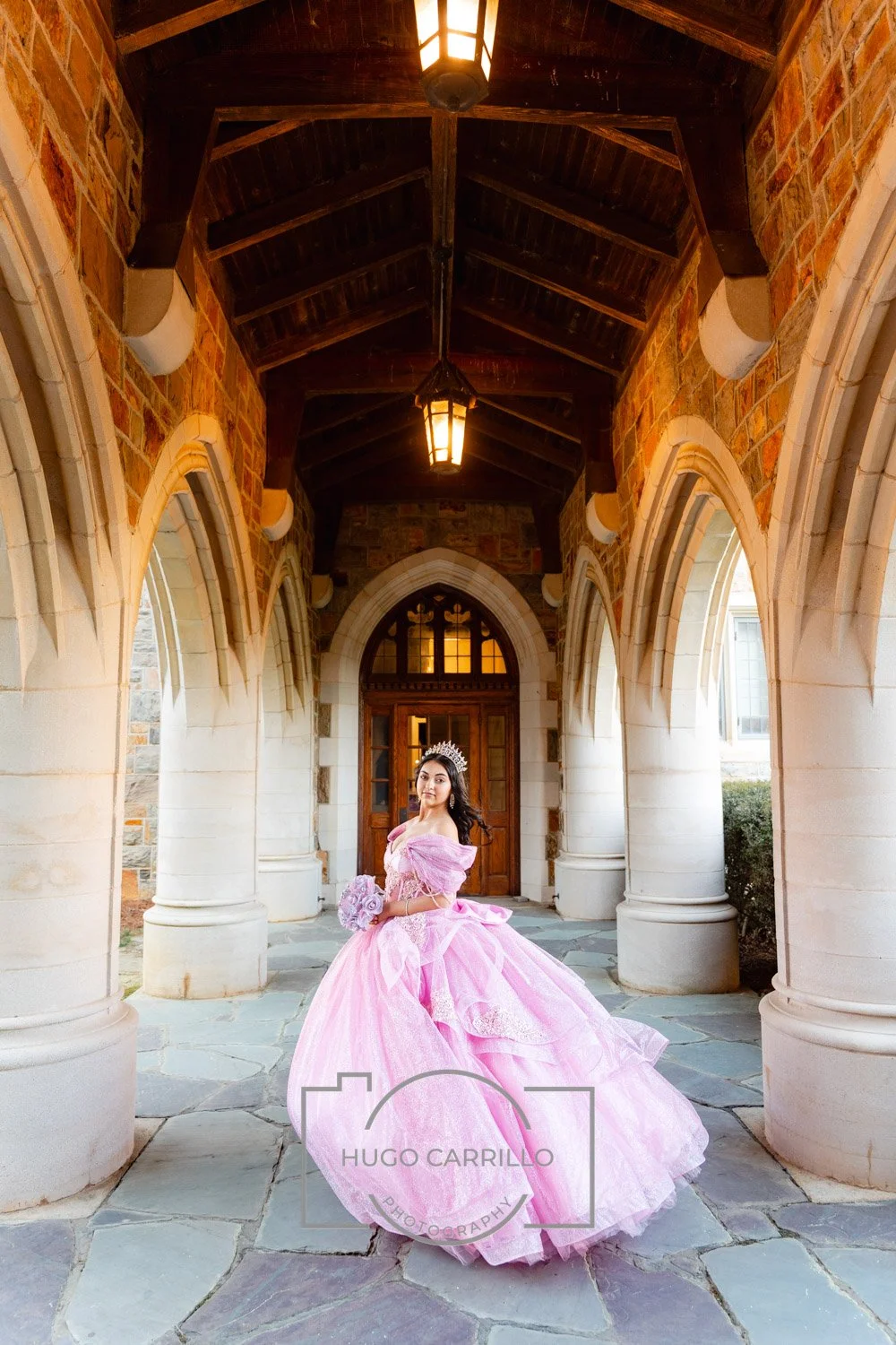 A young woman in a pink princess dress with a tiara standing under a stone archway with wooden roof and hanging lanterns.