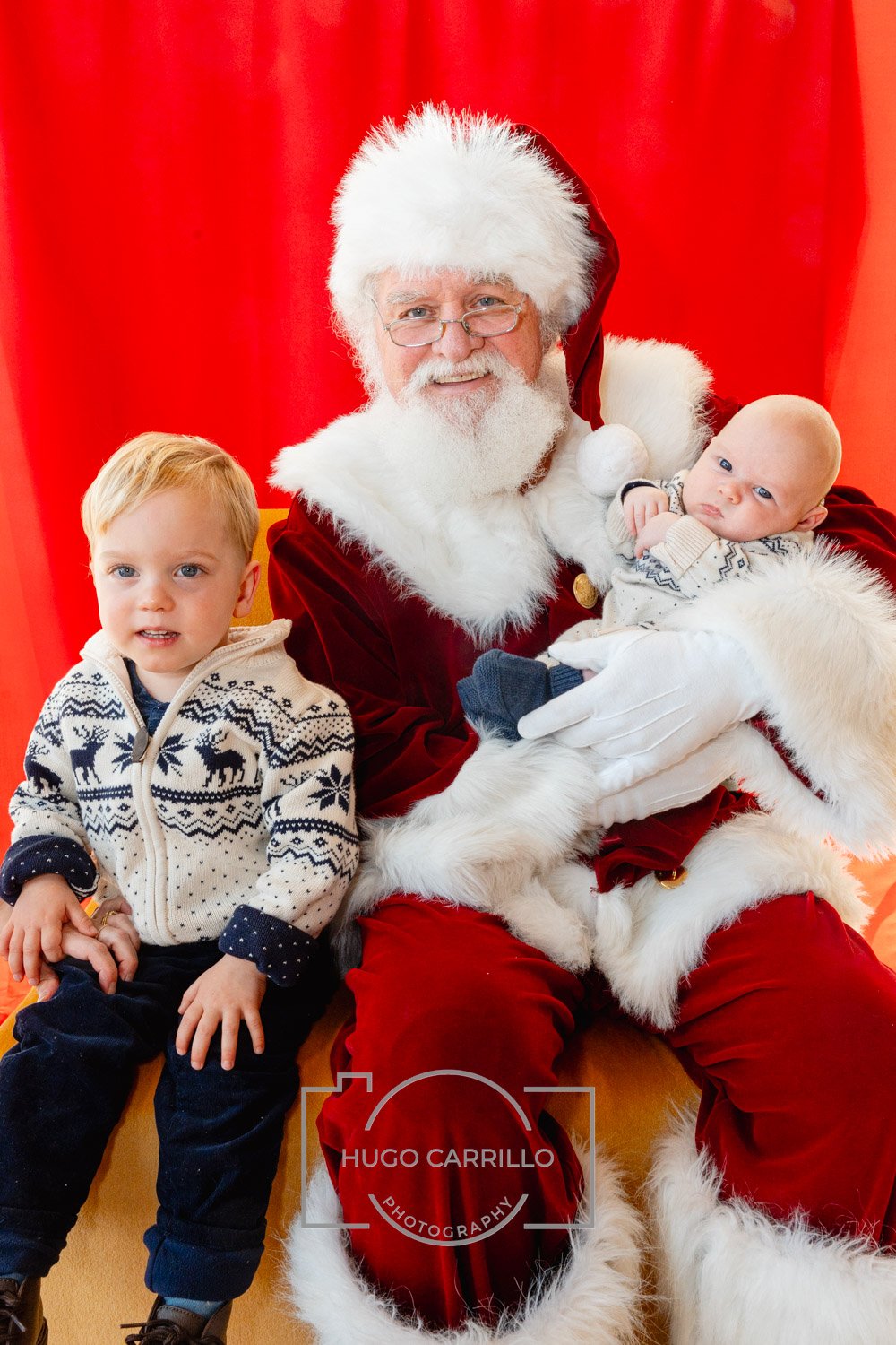 Santa Claus sitting with two young children, one on his lap and the other standing beside him, in front of a red backdrop.