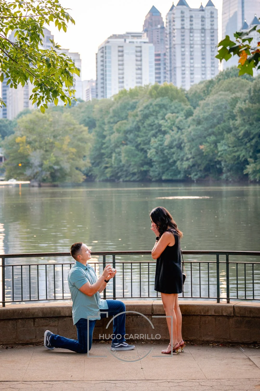 A man kneeling on one knee proposing to a woman by a lake in a park, with city buildings in the background.