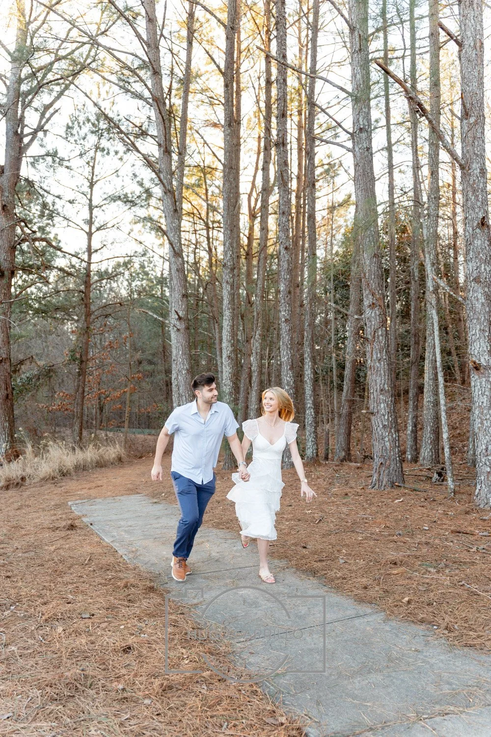 A couple running hand in hand on a forest trail during sunset, surrounded by tall trees and dry grass.