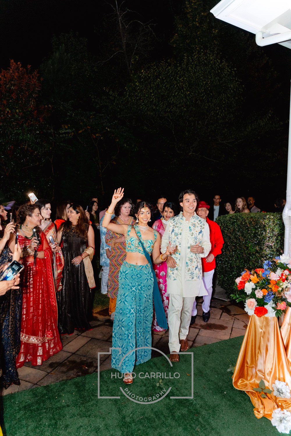 Group of people attending a celebration outdoors at night, with some dressed in traditional South Asian attire, standing near a table decorated with flowers.