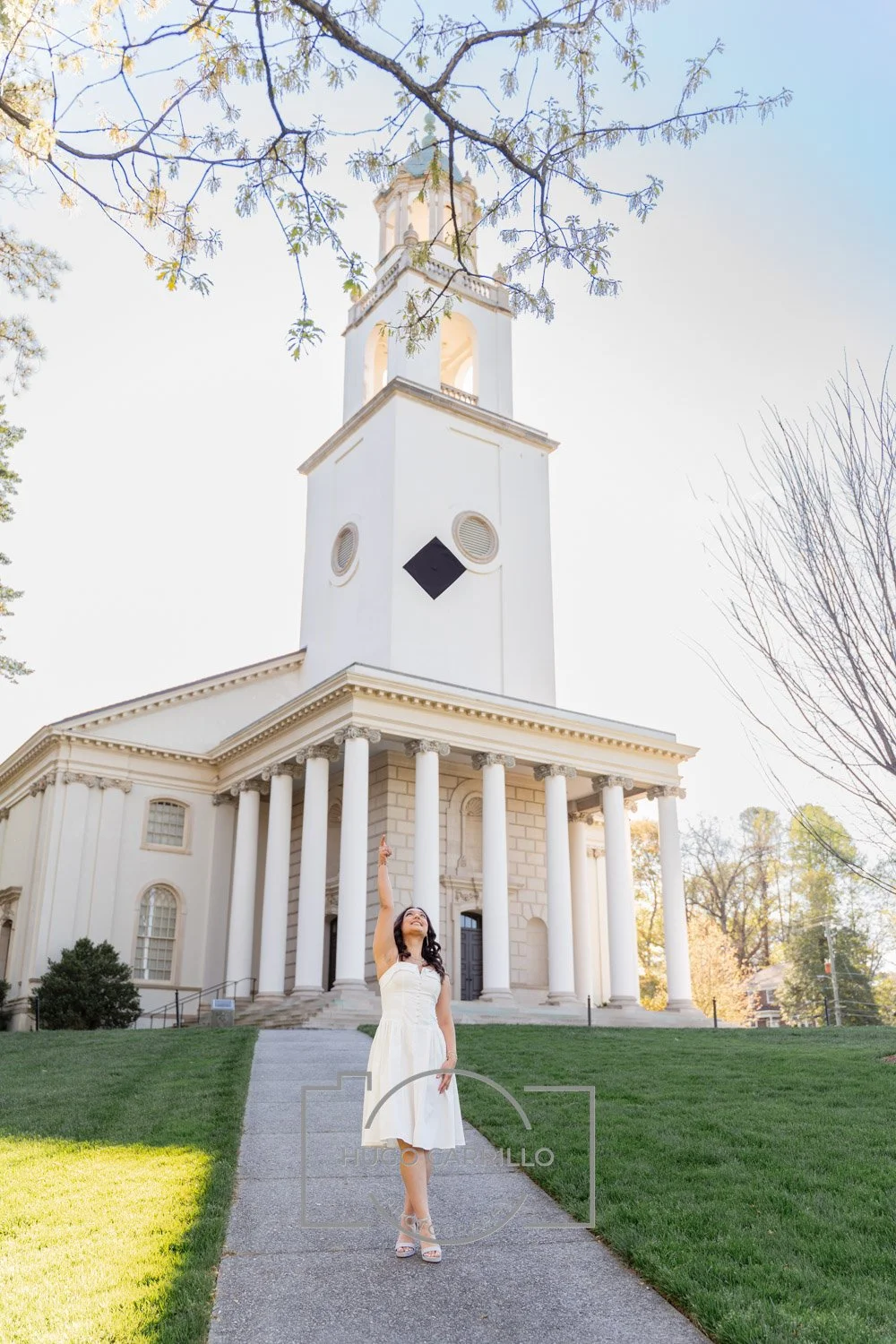 A woman in a white dress standing on a sidewalk, pointing upwards, with a large white church with tall columns in the background on a sunny day.