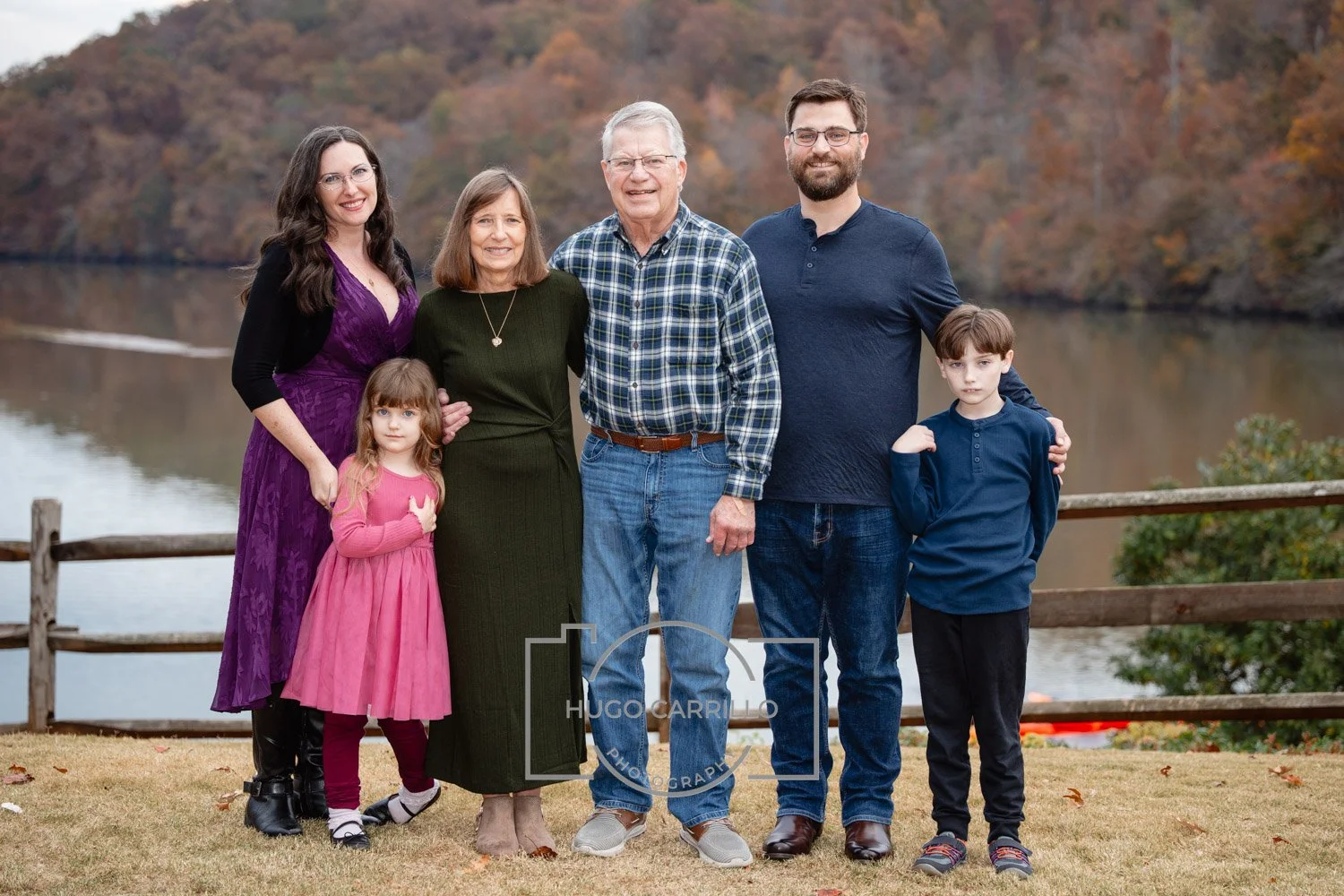 Family of seven outdoors by a river with fall foliage in the background, standing on grass and smiling at the camera.