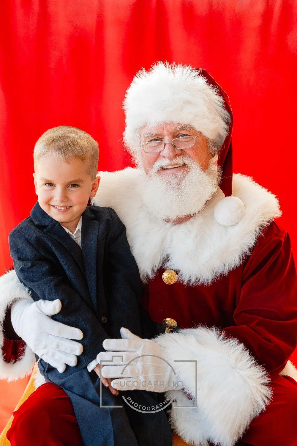 Smiling young boy in a dark suit sitting on Santa Claus's lap. Santa is wearing a traditional red and white suit with glasses, against a red curtain background.