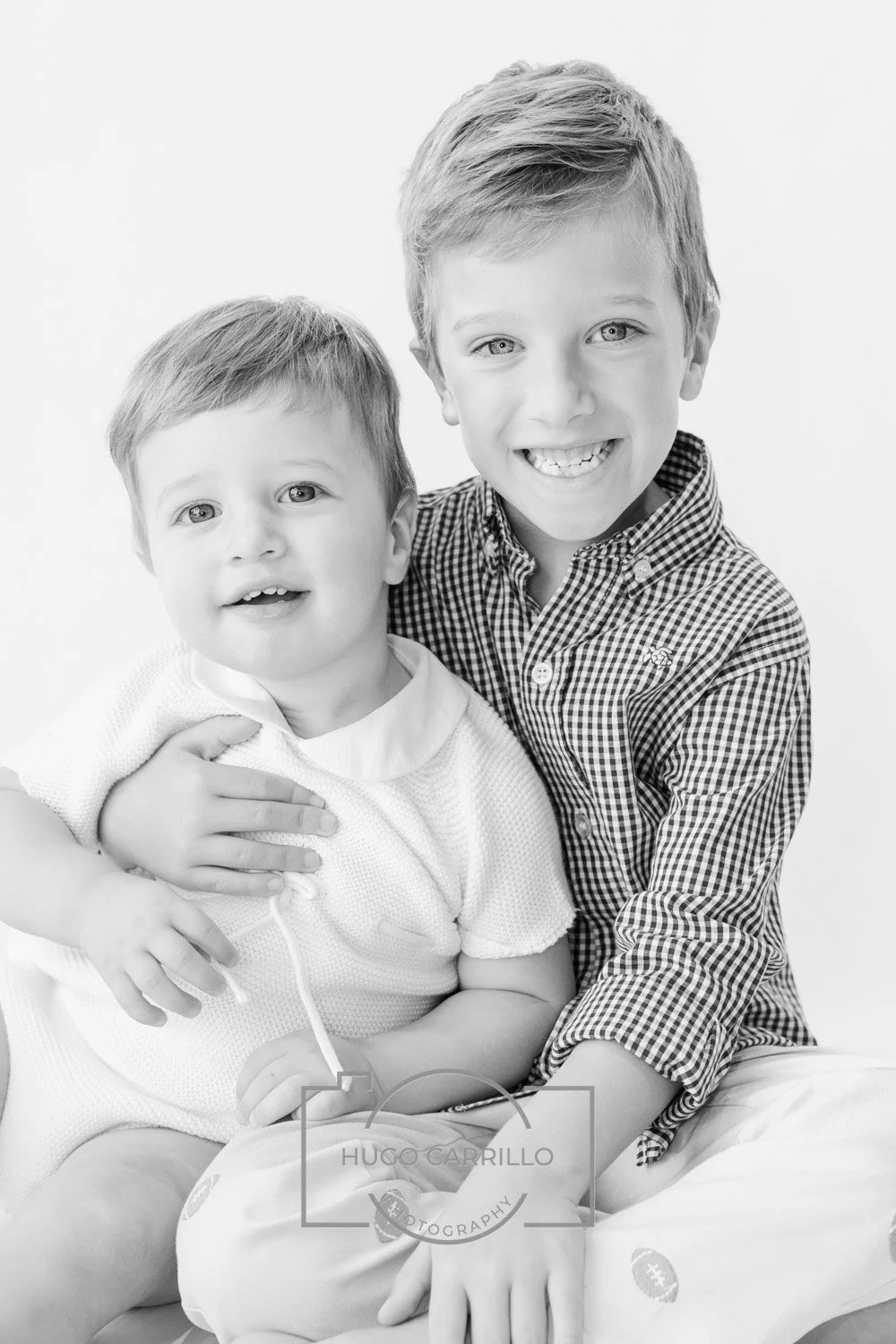Two young boys smiling, one older and one younger, sitting close together against a white background.
