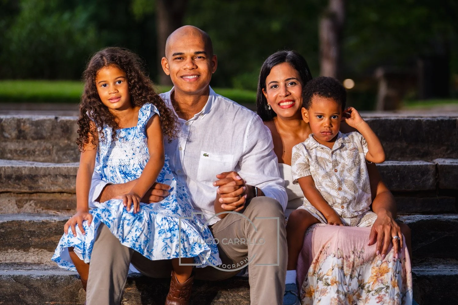 A family of five sitting outdoors on stone steps, smiling at the camera, with trees in the background.