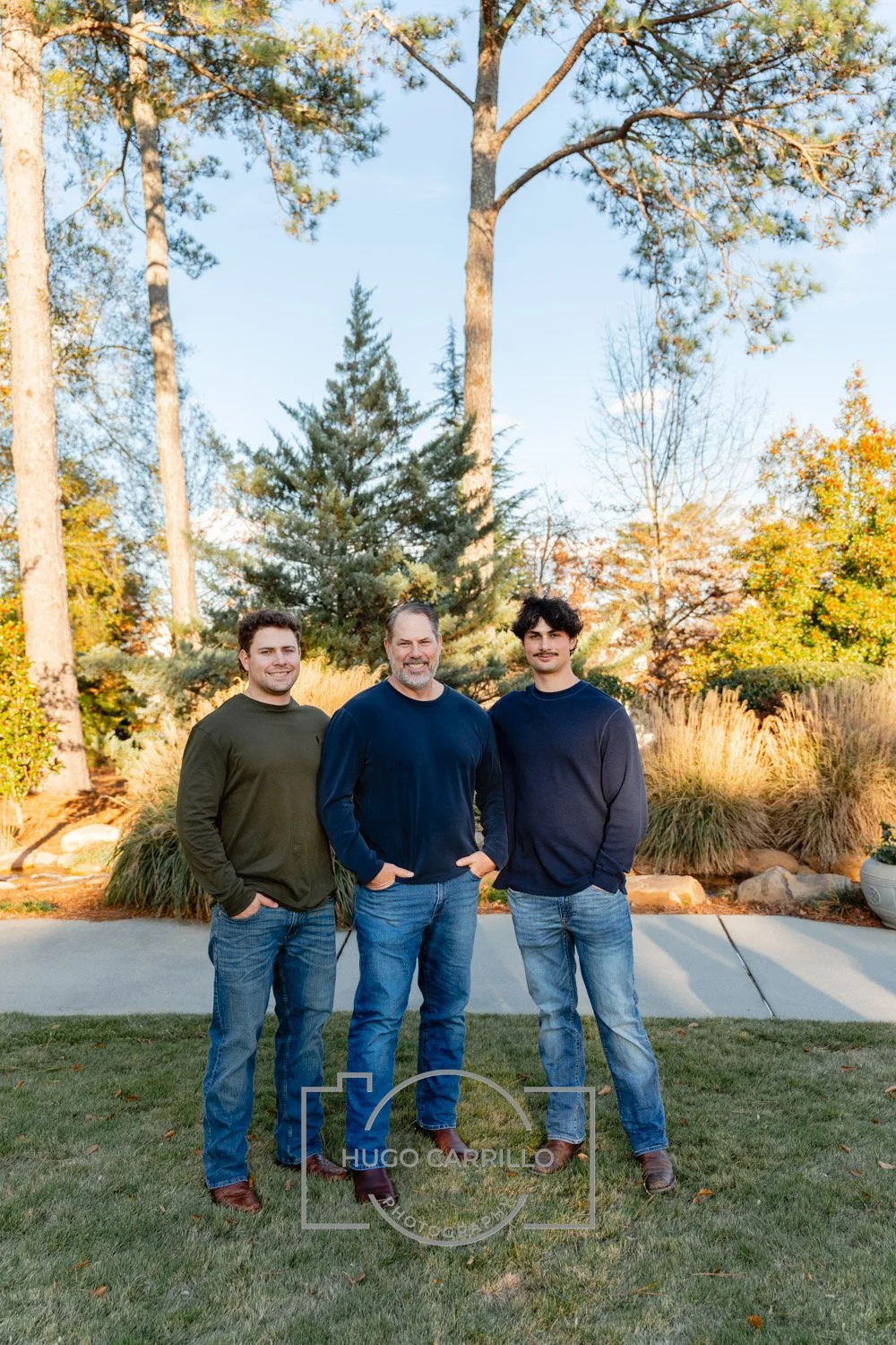 Three men standing outdoors in a park-like setting with trees and bushes in the background, smiling and posing for a photo.