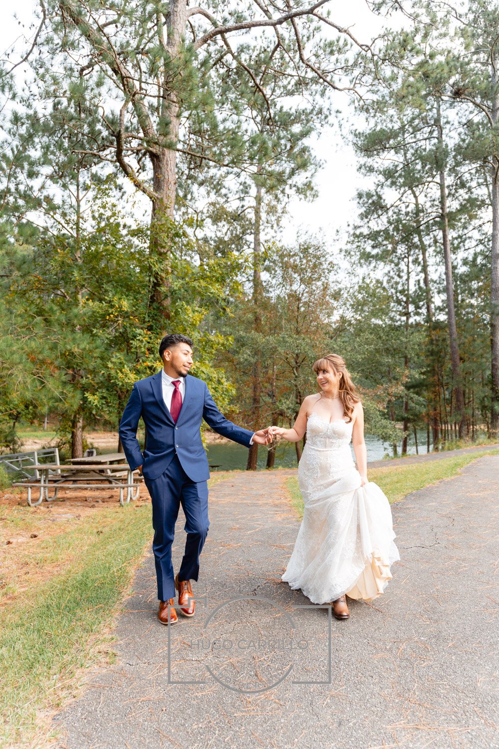 A bride and groom holding hands and walking in a park surrounded by tall trees and a lake in the background.
