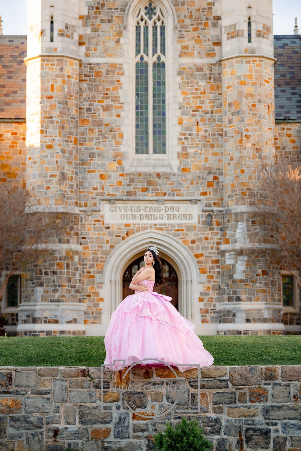 A quinceañera in a pink ball gown and tiara standing in front of a stone church with stained glass windows. The church has a sign that says, "GIVE US THIS DAY OUR DAILY BREAD."