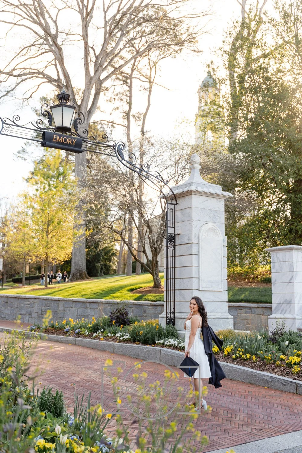 A young woman in a white dress holding a black umbrella and a black coat over her shoulder, standing on a brick sidewalk near a stone archway with the word "Emory" on it, in a park-like setting with trees, flowers, and a lawn, during late afternoon o