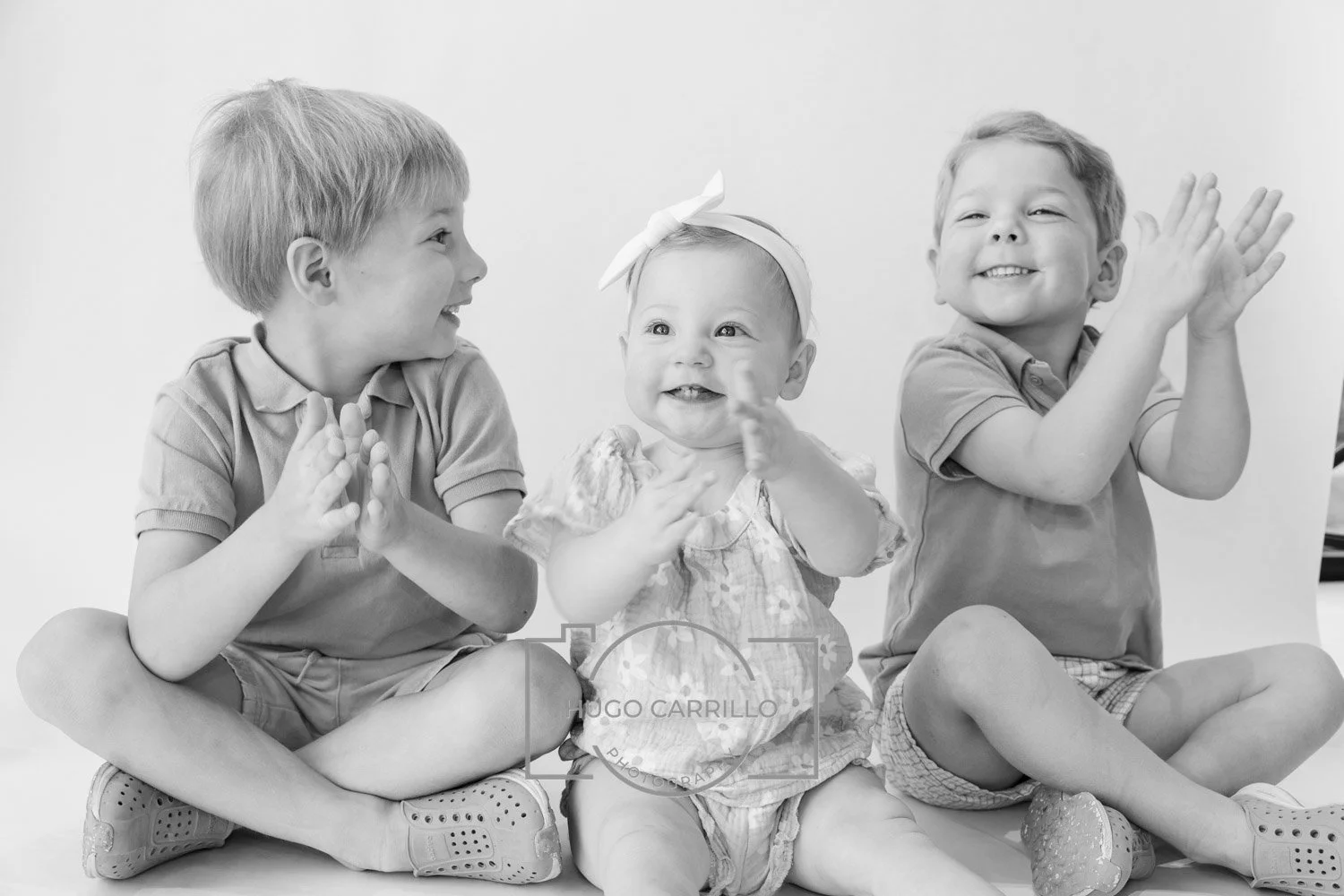 Three young children, two boys and one girl, sitting cross-legged and clapping hands, smiling and looking at each other in a joyful moment.