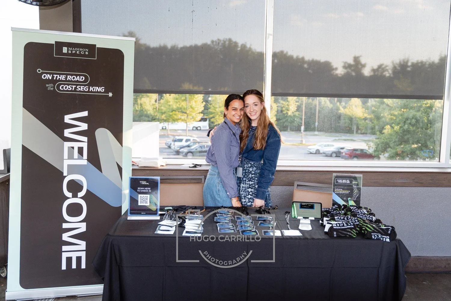 Two women smiling and hugging at a registration table for a Madison Specs event, with sunglasses and promotional materials displayed on the table, and a large Madison Specs sign to the side.