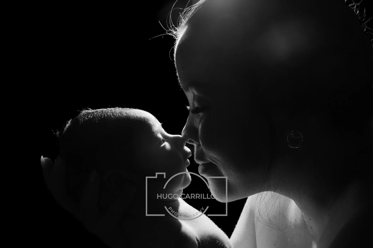 Black and white photo of a woman gently holding a baby close, with their faces touching and eyes closed, illuminated from behind creating a silhouette effect.