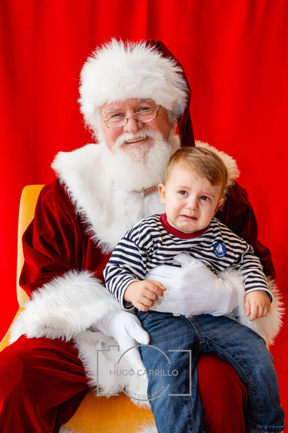 Santa Claus with a white beard, glasses, and a red suit, sitting on a chair, holding a young boy who is making a worried or confused expression. The background is red.