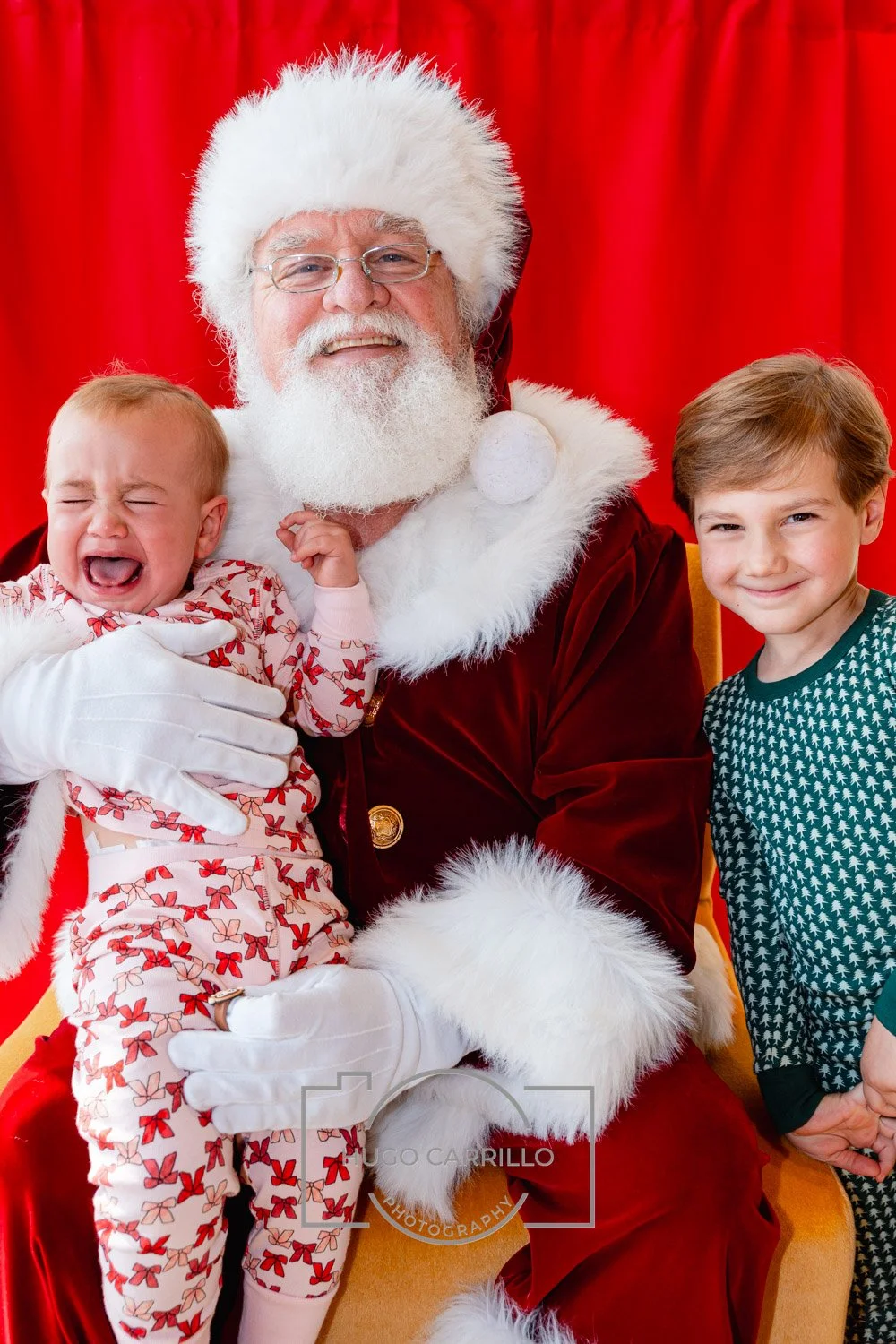 Santa Claus with two children on his lap against a red backdrop; one child is crying, and the other smiling.
