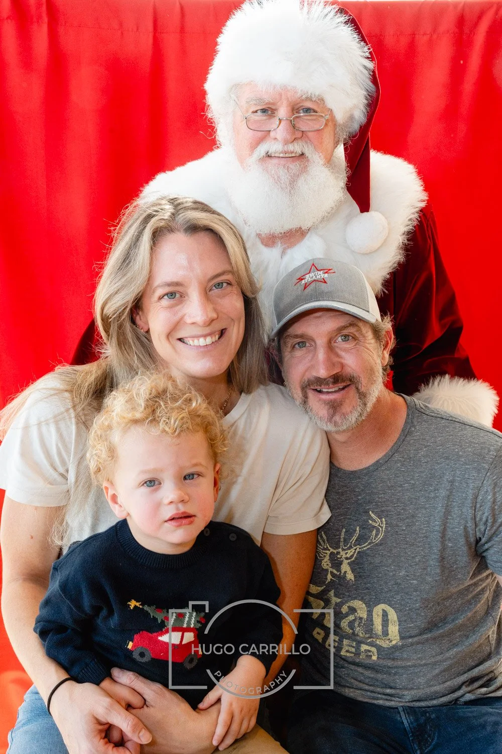 Family photo with a woman, a man, a young boy, and Santa Claus, in front of a red backdrop.