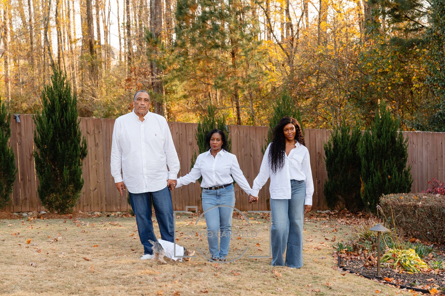 Three people holding hands and standing in a backyard with trees in the background and a wooden fence, a small dog on a leash, autumn leaves on the ground, and small shrubs and garden decor.