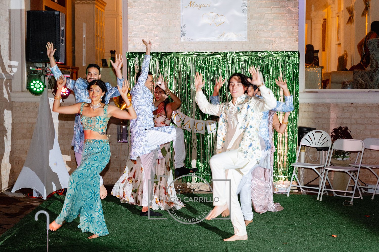 People dancing and celebrating at a wedding reception with a green metallic fringe backdrop and a banner that says 'CHARGEE'.