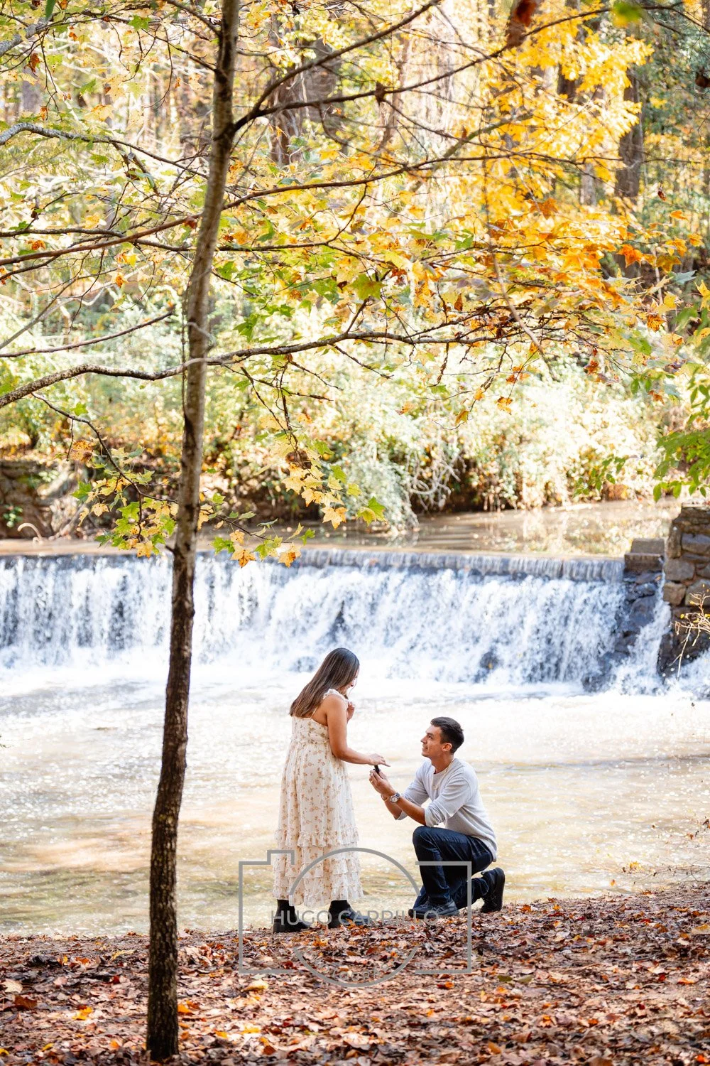 A man proposes marriage to a woman by a waterfall in a forest with autumn leaves.
