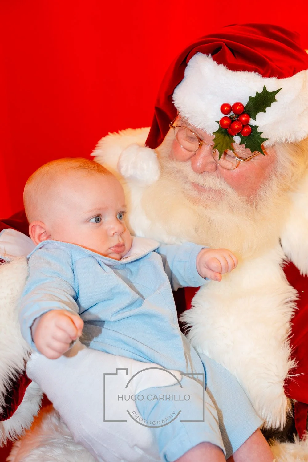 A young child sitting on Santa Claus's lap, wearing a light blue shirt, looking at Santa with a curious expression. Santa is dressed in his traditional red suit with white fur trim, wearing glasses, and has a holly decoration on his hat. The backgrou