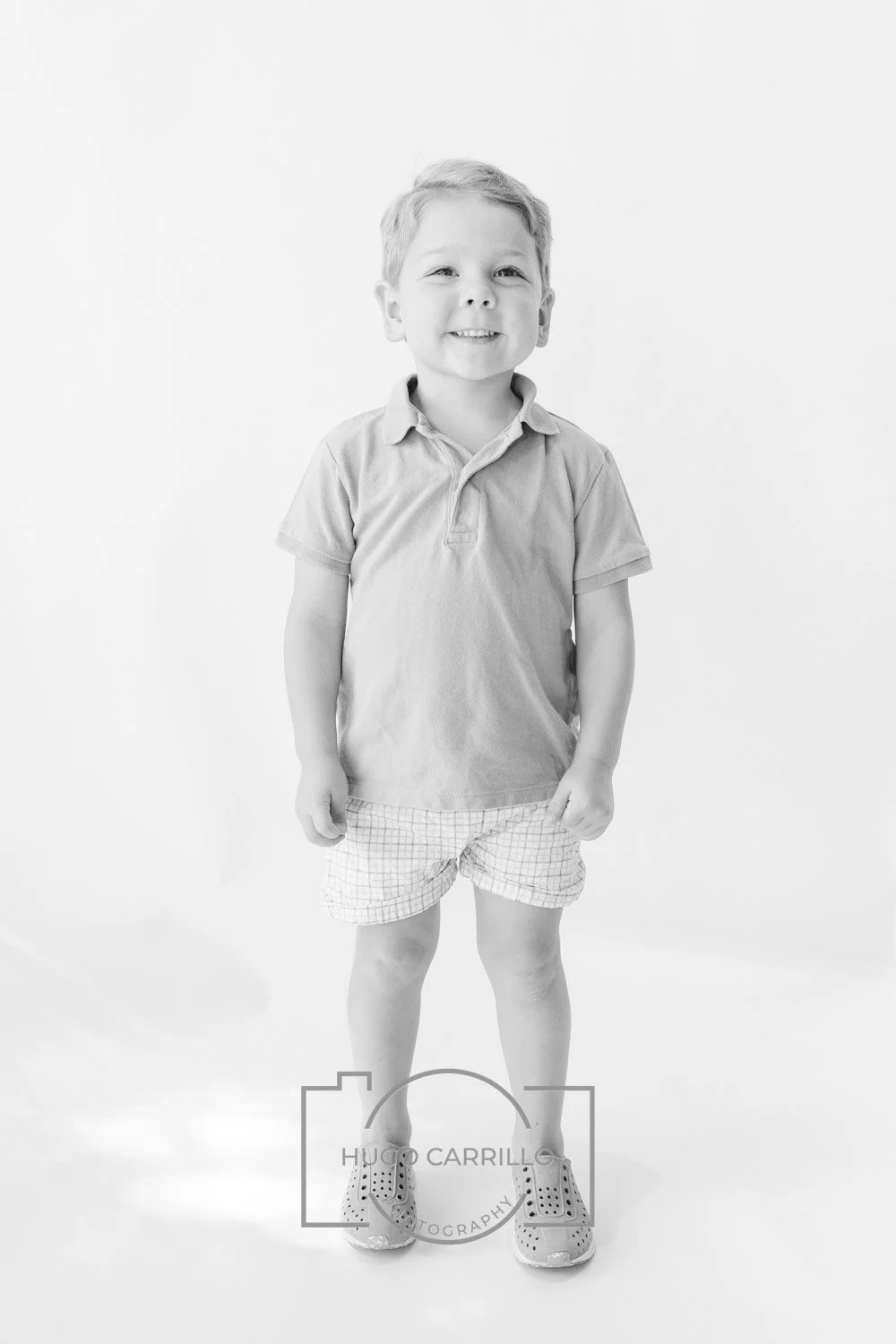 A young boy smiling, standing against a plain background. He is wearing a short-sleeved polo shirt, shorts, and sneakers.