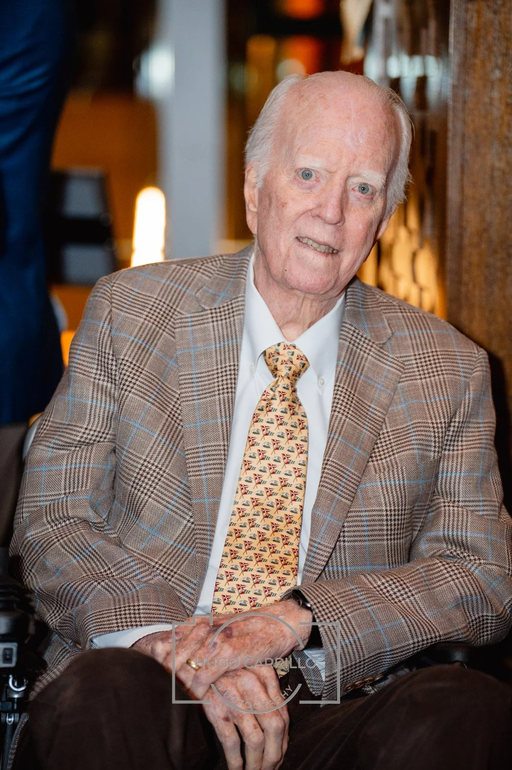 An elderly man with white hair and blue eyes, wearing a beige checked blazer, a white shirt, and a patterned tie, sitting indoors in a restaurant or event space.