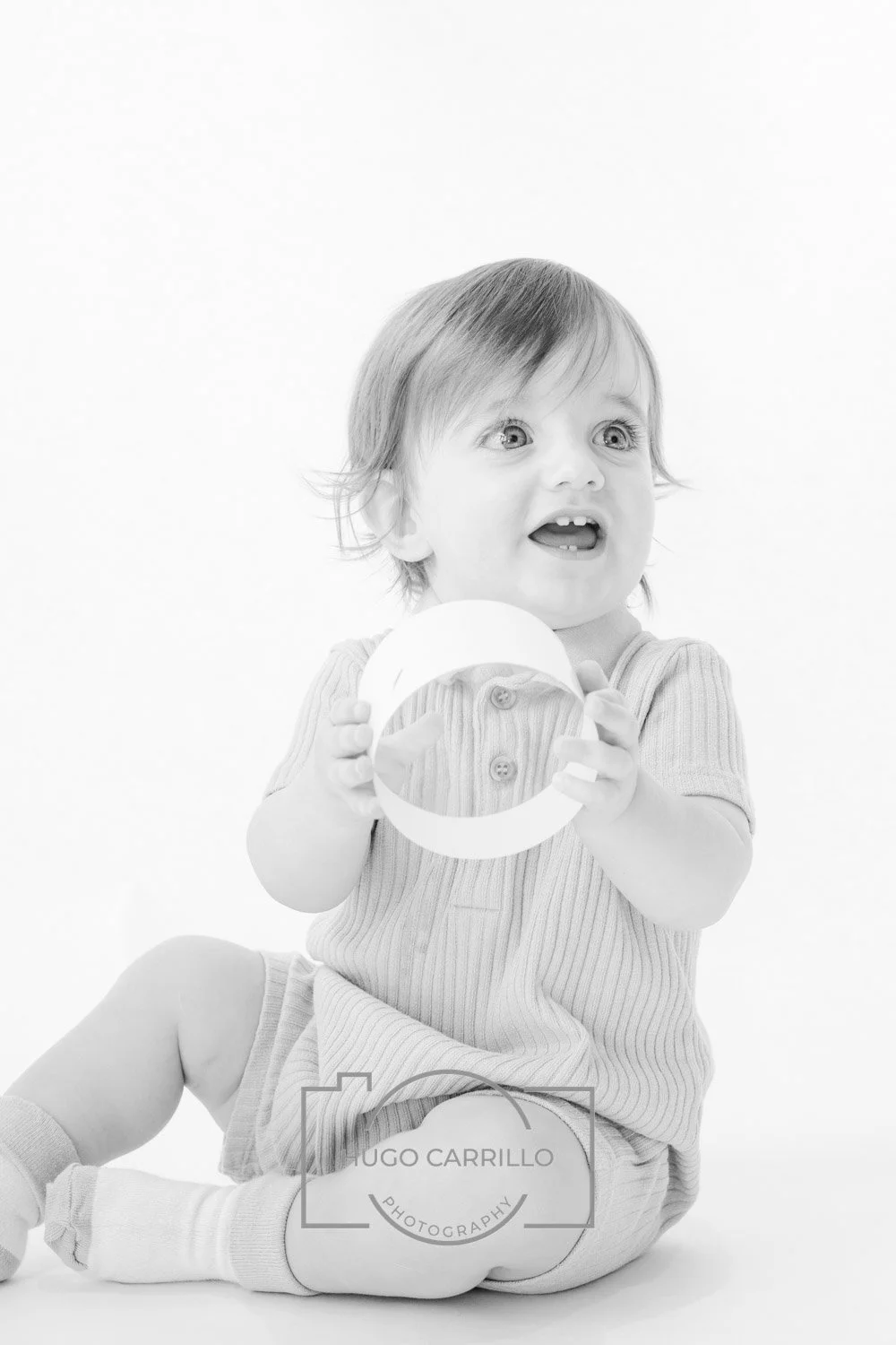 A young child with light-colored hair sitting on the floor, holding a circular object and looking with an expression of surprise or curiosity, against a plain white background.