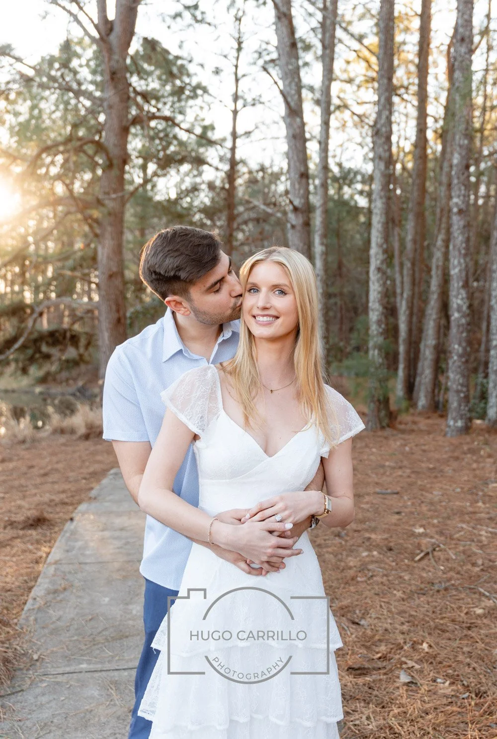 A man is kissing a woman on the cheek in a wooded outdoor setting during sunset. The woman is smiling and wearing a white dress, and the man is dressed in a light blue shirt.