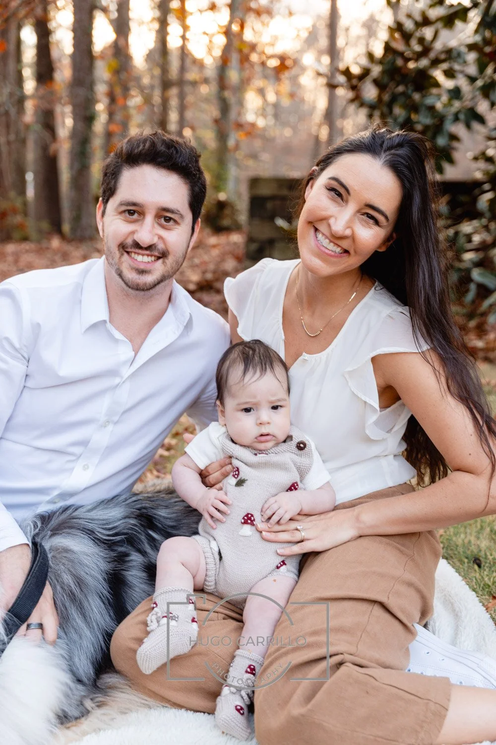 A happy family of three, including a man, a woman, and a baby, sitting outdoors in a wooded area during autumn. The woman is holding the baby, and they are all smiling, with the man and woman wearing white tops and the baby in a beige outfit.