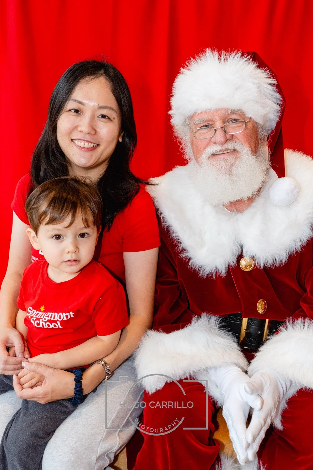 A woman and a young boy sitting next to Santa Claus in front of a red backdrop, all dressed in red shirts, during a Christmas event.