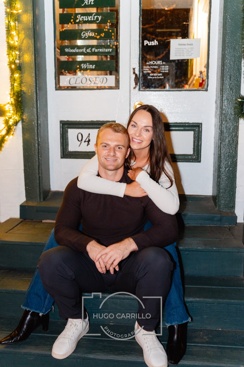 A smiling man and woman sitting together on the steps outside a shop with signs for art, jewelry, gifts, woodworking, wine, and a closed sign, during holiday season with festive lights.