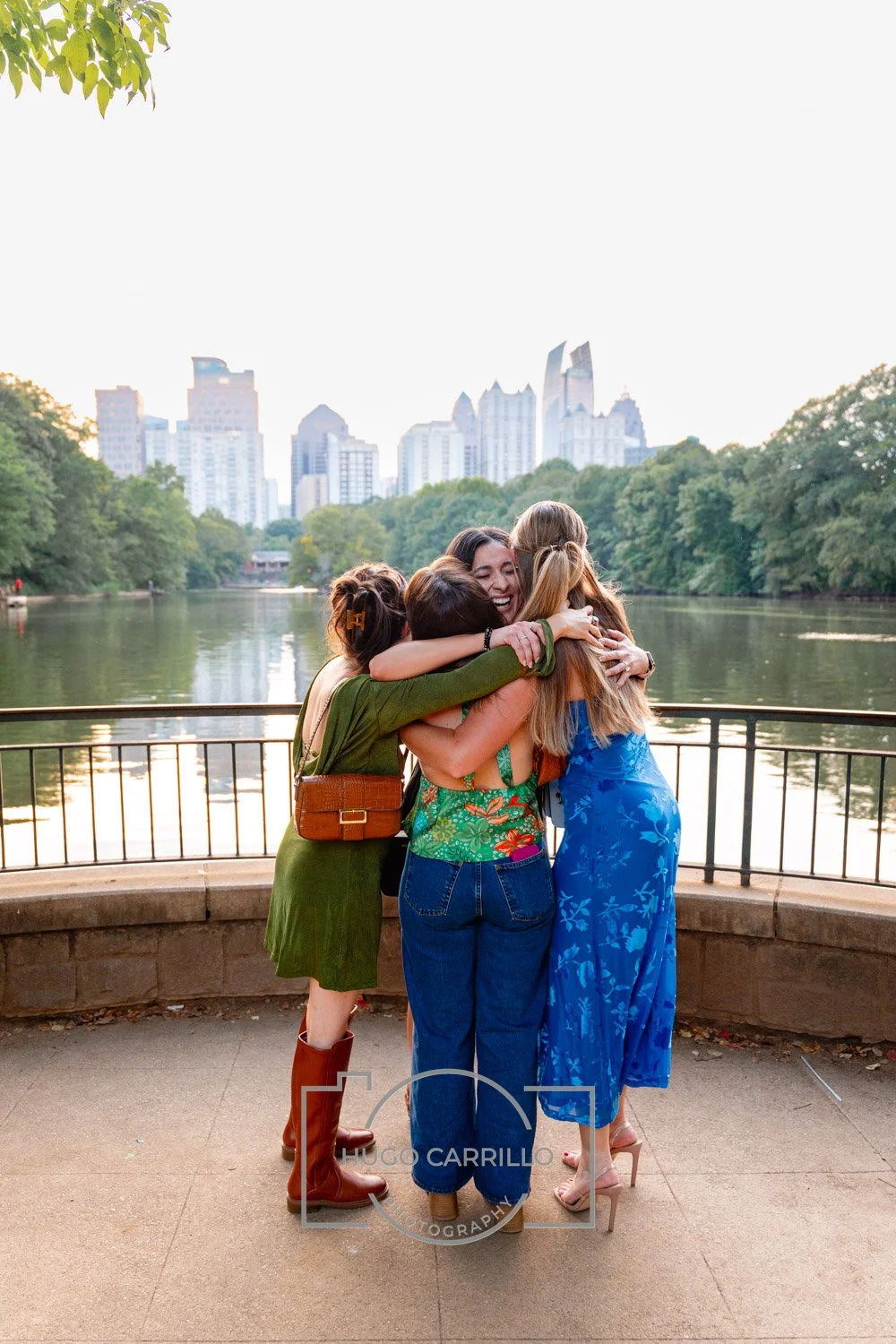 Four women hugging each other near a lake with city skyline in the background during daytime.
