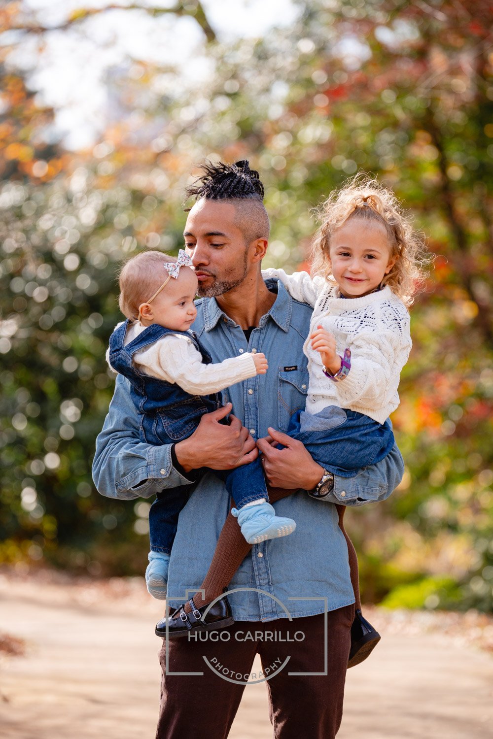 A man holding two young girls outdoors during fall, with colorful autumn leaves in the background. The man is wearing a blue denim shirt, and one girl is in a white sweater and has curly blonde hair, while the other girl, held close to his face, has 