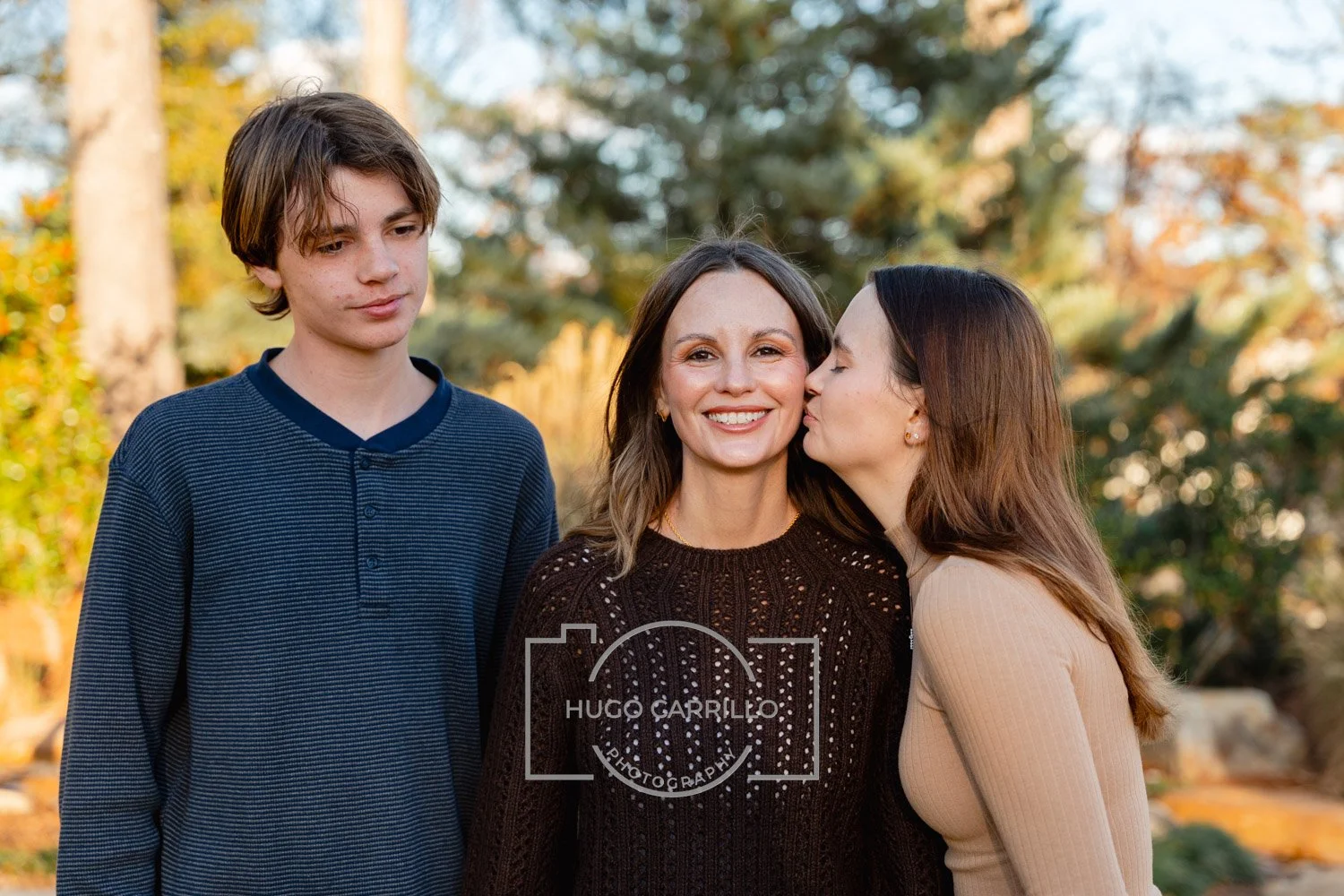 A woman smiling as she is kissed on the cheek by another woman, with a teenage boy standing nearby, outdoors with autumn foliage in the background.