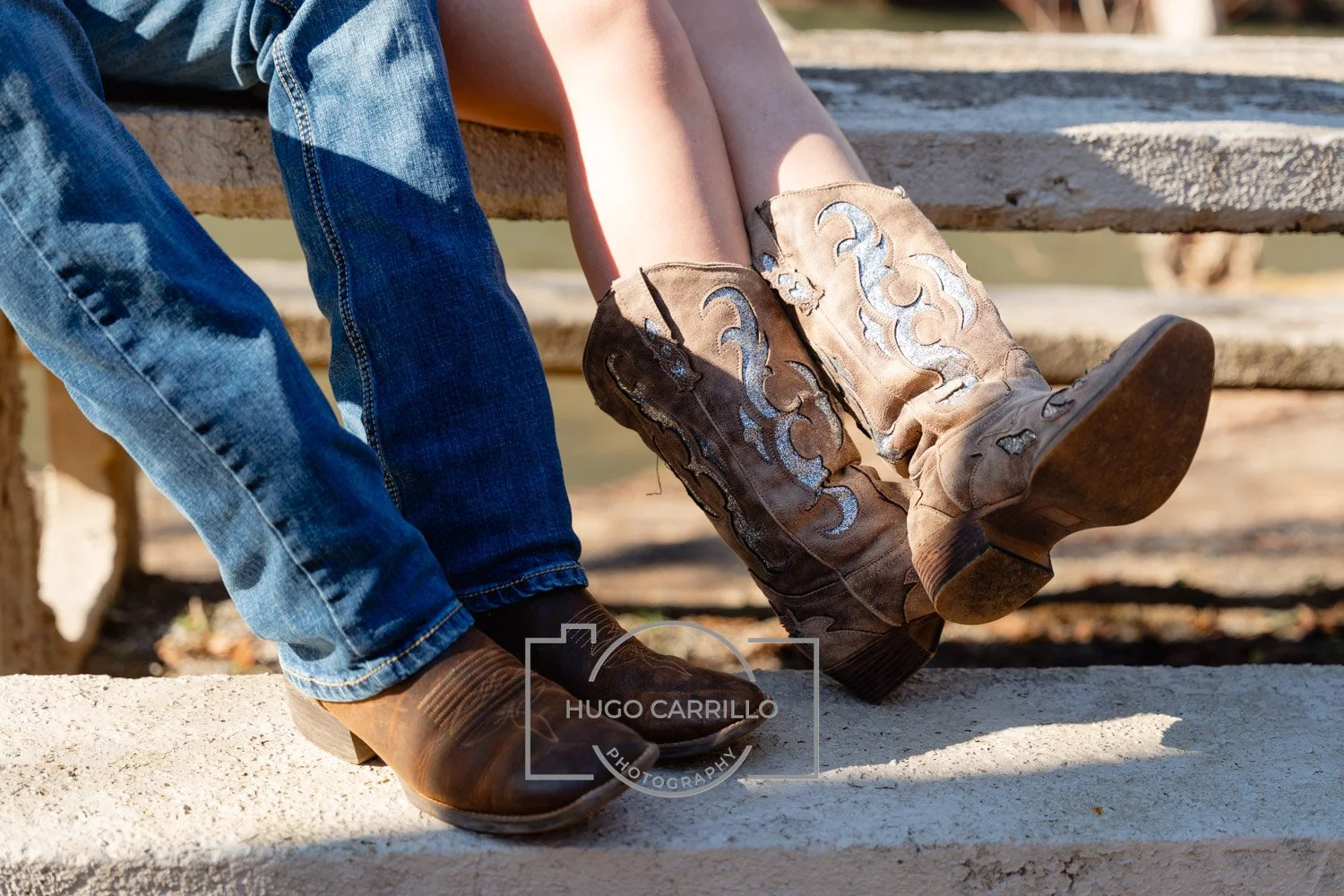 Close-up of a person wearing cowboy boots sitting on a wooden bench with another person's legs visible next to them, both outdoors.
