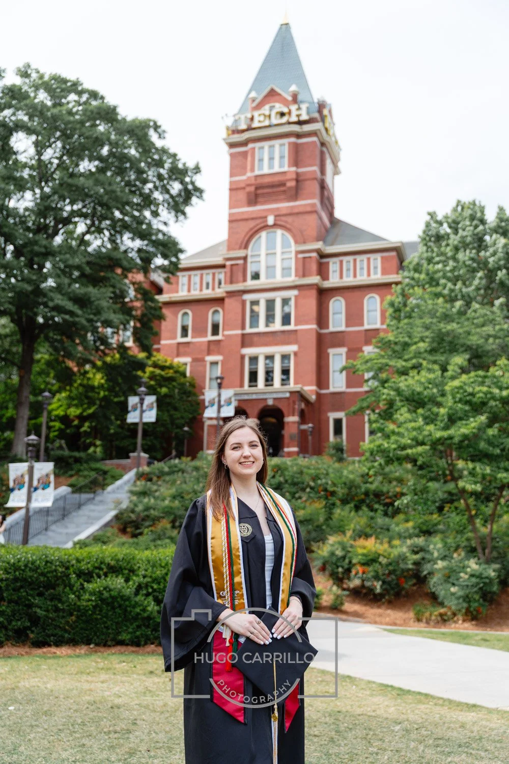 A young woman in graduation cap and gown standing outdoors in front of a historic campus building with a tall clock tower surrounded by trees and greenery.
