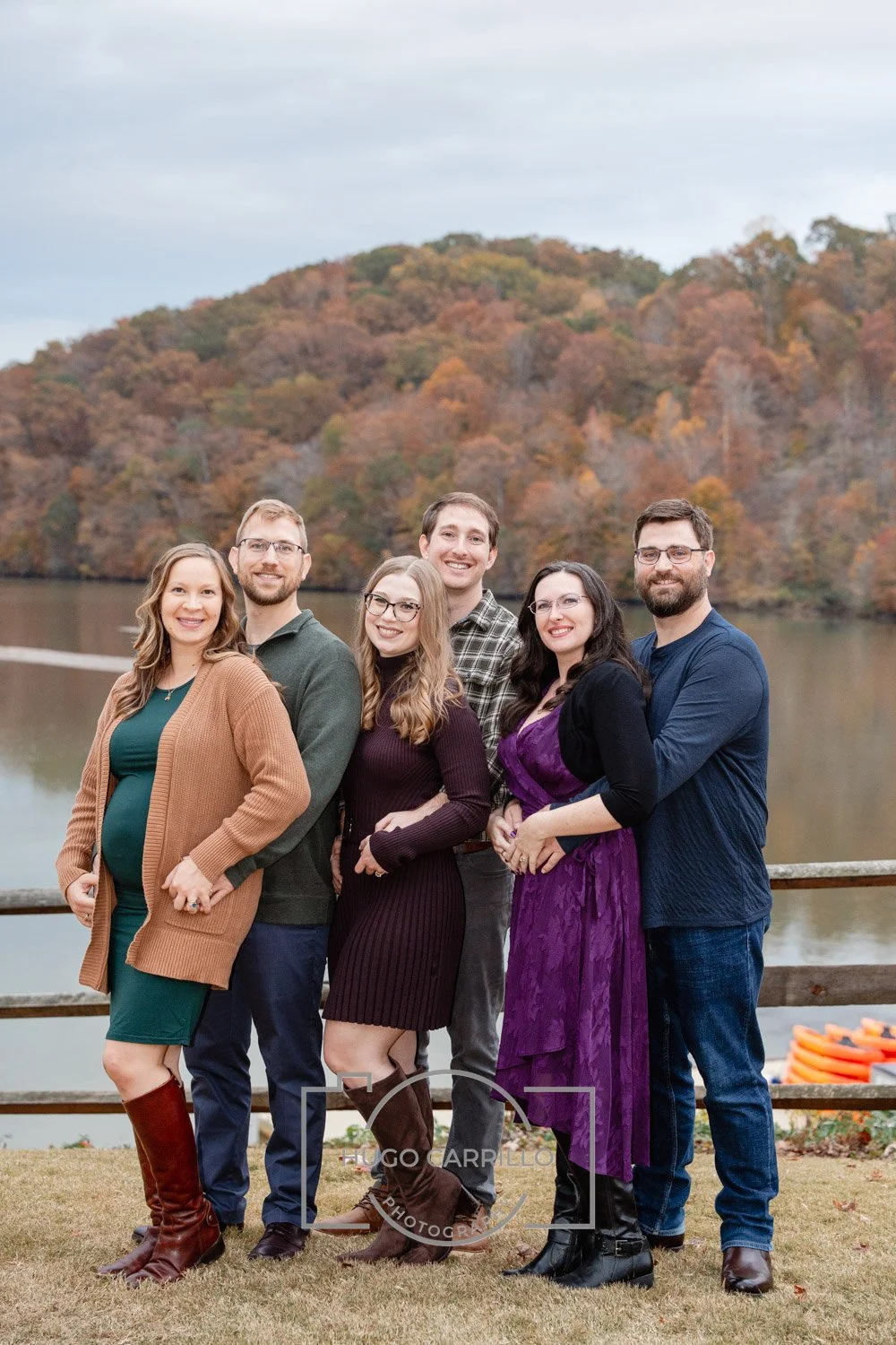 Group of six people standing outdoors near a lake, with fall foliage in the background, smiling at the camera.