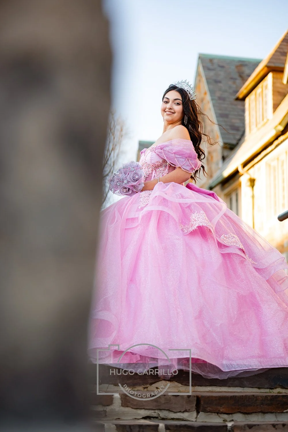 A quinceañera wearing a pink princess gown and tiara, smiling outdoors with houses in the background.