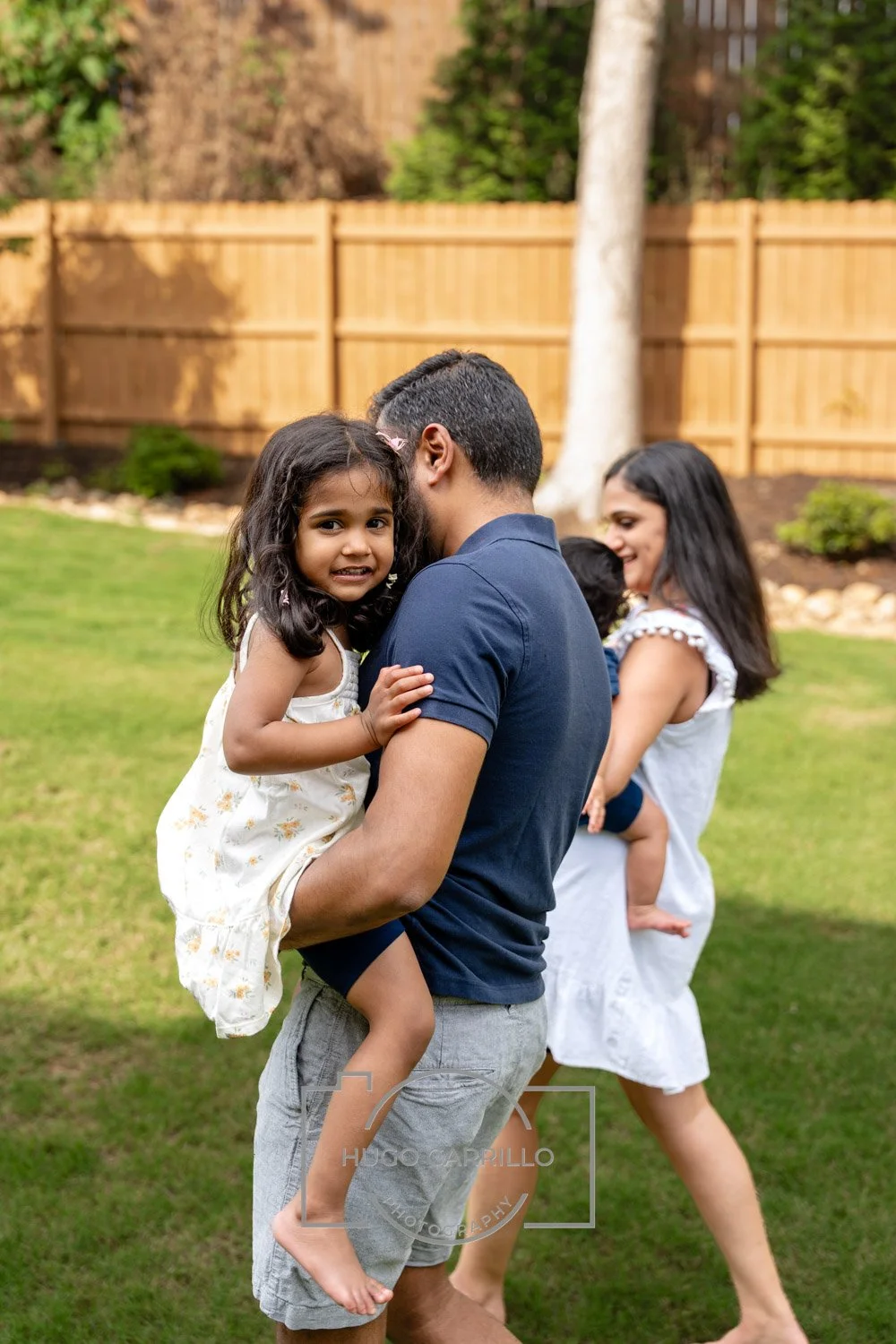 A man holding a young girl in a backyard, with a woman walking behind them, both smiling. The girl looks slightly apprehensive. The background features a wooden fence, trees, and green grass.
