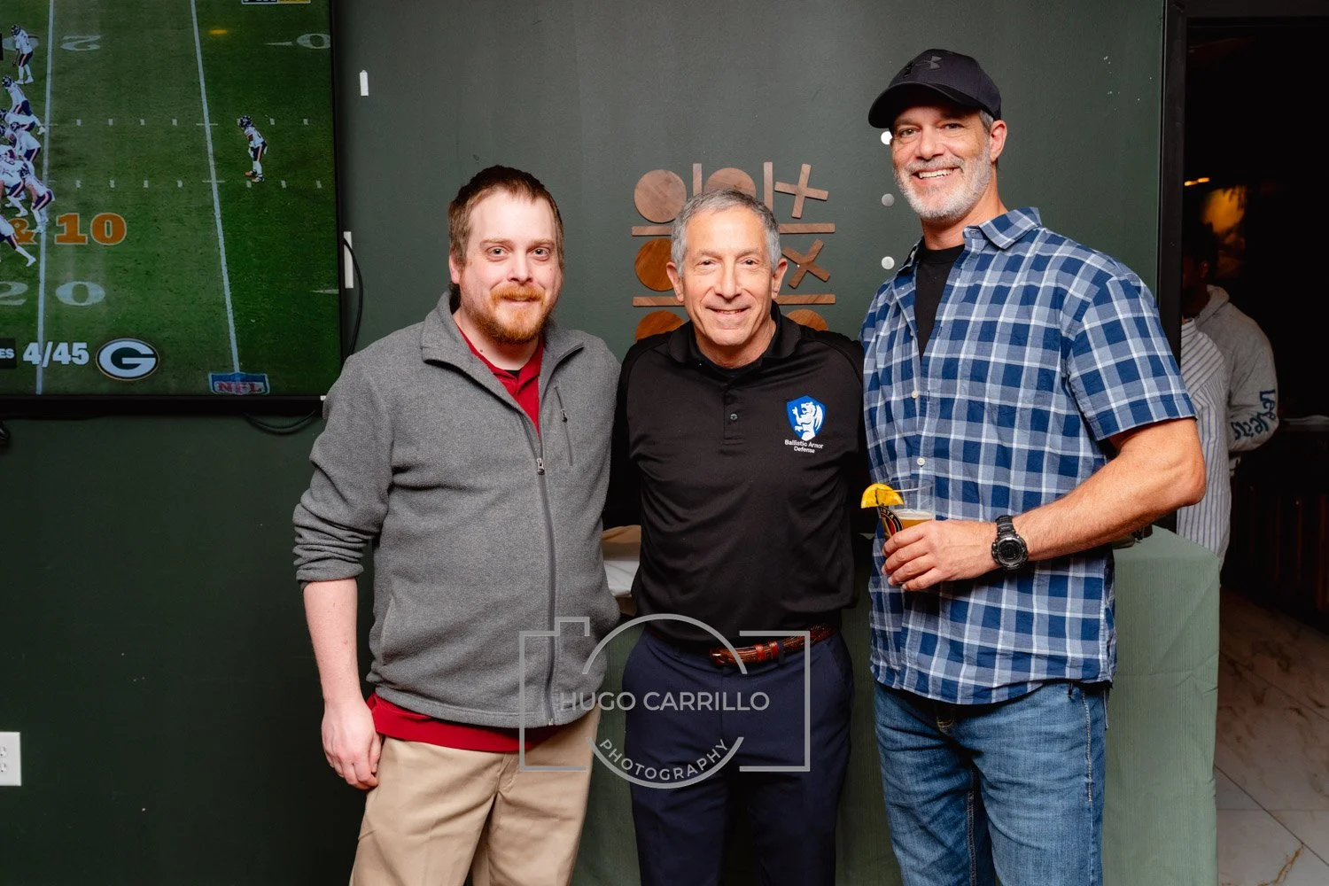 Three men standing close together indoors, smiling at the camera; the man on the right holds a drink with a lemon garnish. A TV screen showing a football game is in the background.