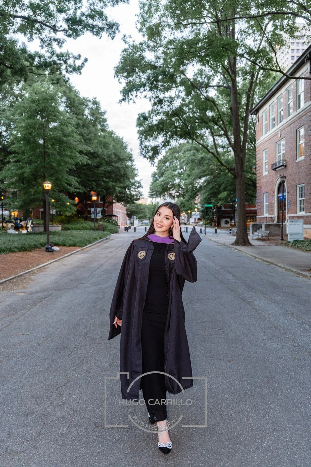 A young woman in a graduation cap and gown standing on a city street with trees and brick buildings, smiling at the camera.