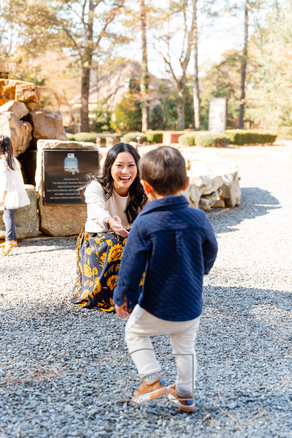 A woman kneeling and smiling while holding hands with a young boy outdoors on a sunny day, with trees and a stone monument in the background.
