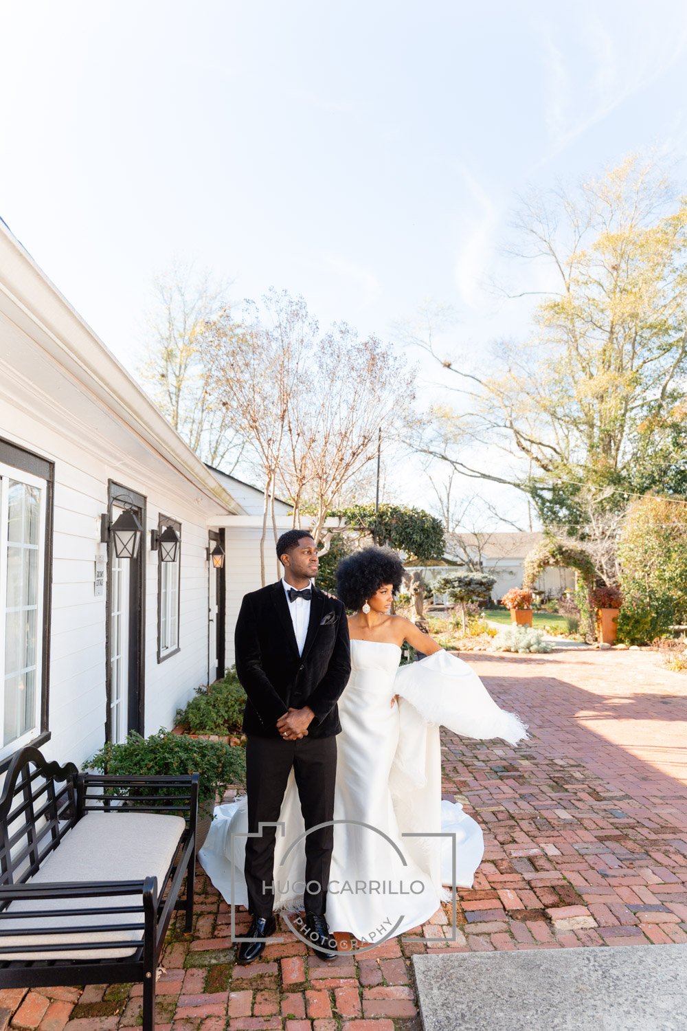 A bride and groom standing outdoors on a brick patio during daytime, with trees and a white house in the background. The bride wears a strapless white wedding gown, and the groom is in a black tuxedo with a bow tie.
