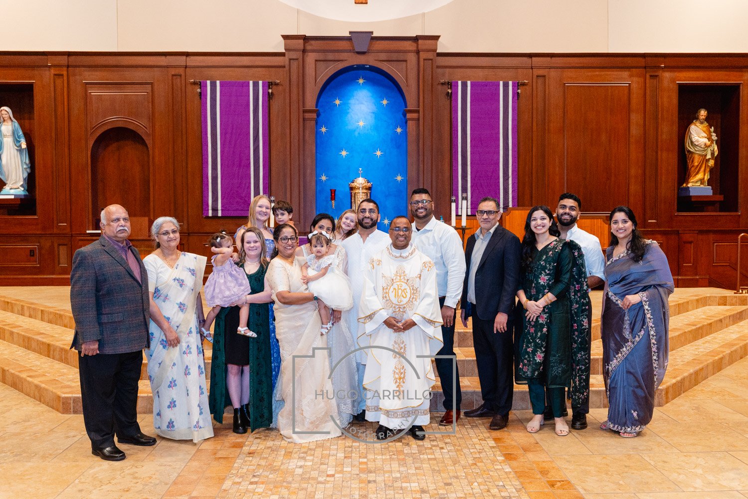 Group of people celebrating a religious ceremony inside a church, standing in front of altar with purple banners, wooden paneling, and statues, with the priest in white robes at the center.